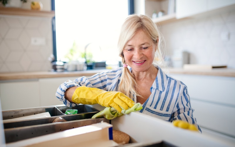 Before photo of a cluttered under-sink cabinet with cleaning supplies, plastic bags, and sponges crammed together haphazardly